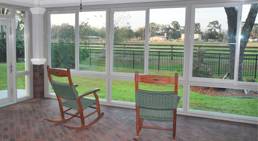two wood rocking chairs sitting inside a glass sunroom looking out to a backyard